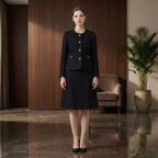 Woman in a black outfit standing in a modern interior setting with a plant and chair.