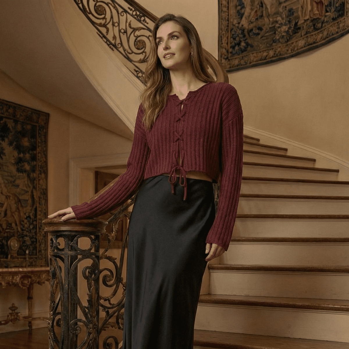 Woman in a maroon top and dark skirt standing on a staircase in an elegant interior setting.