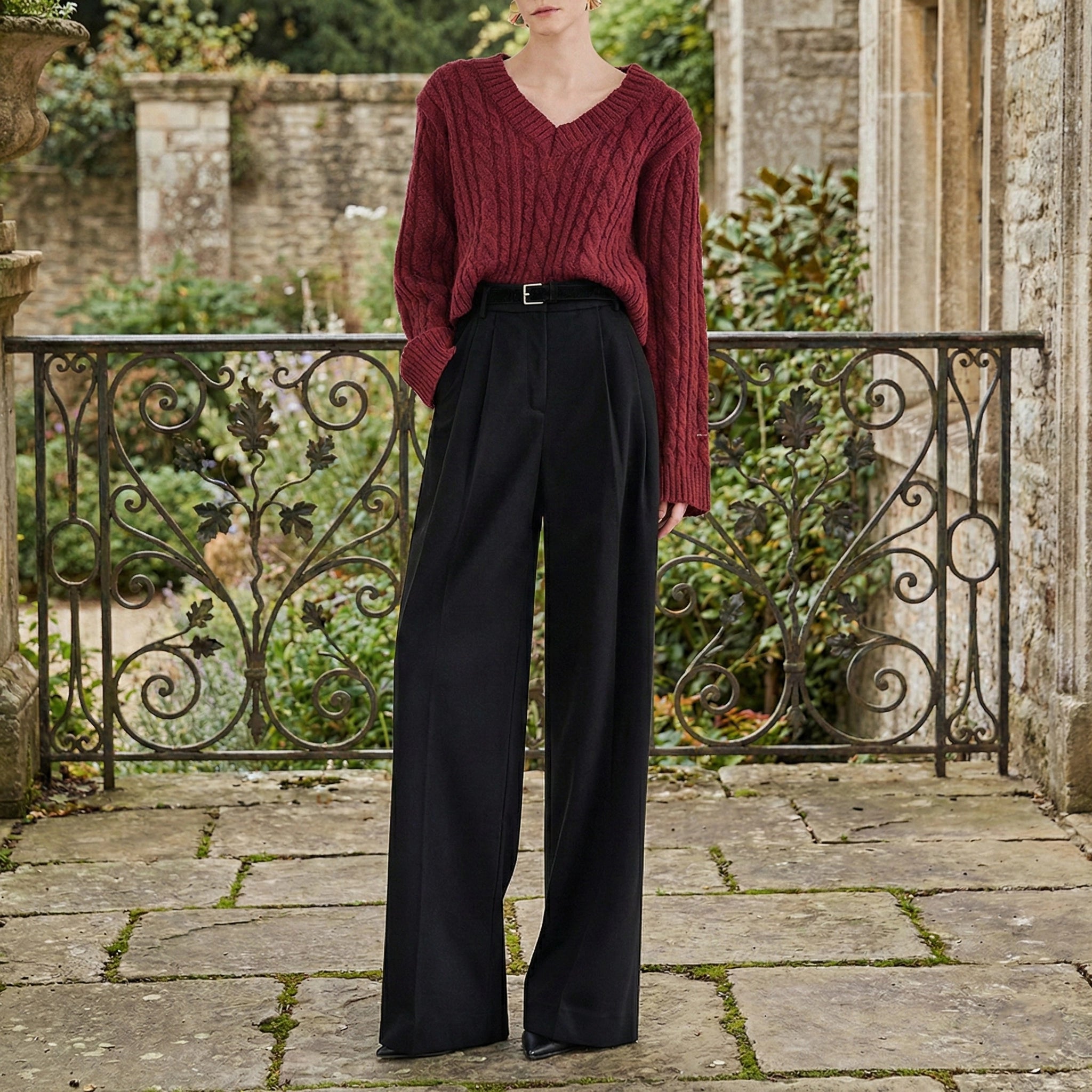 Person wearing a red sweater and black pants standing on a stone patio with garden and building in the background