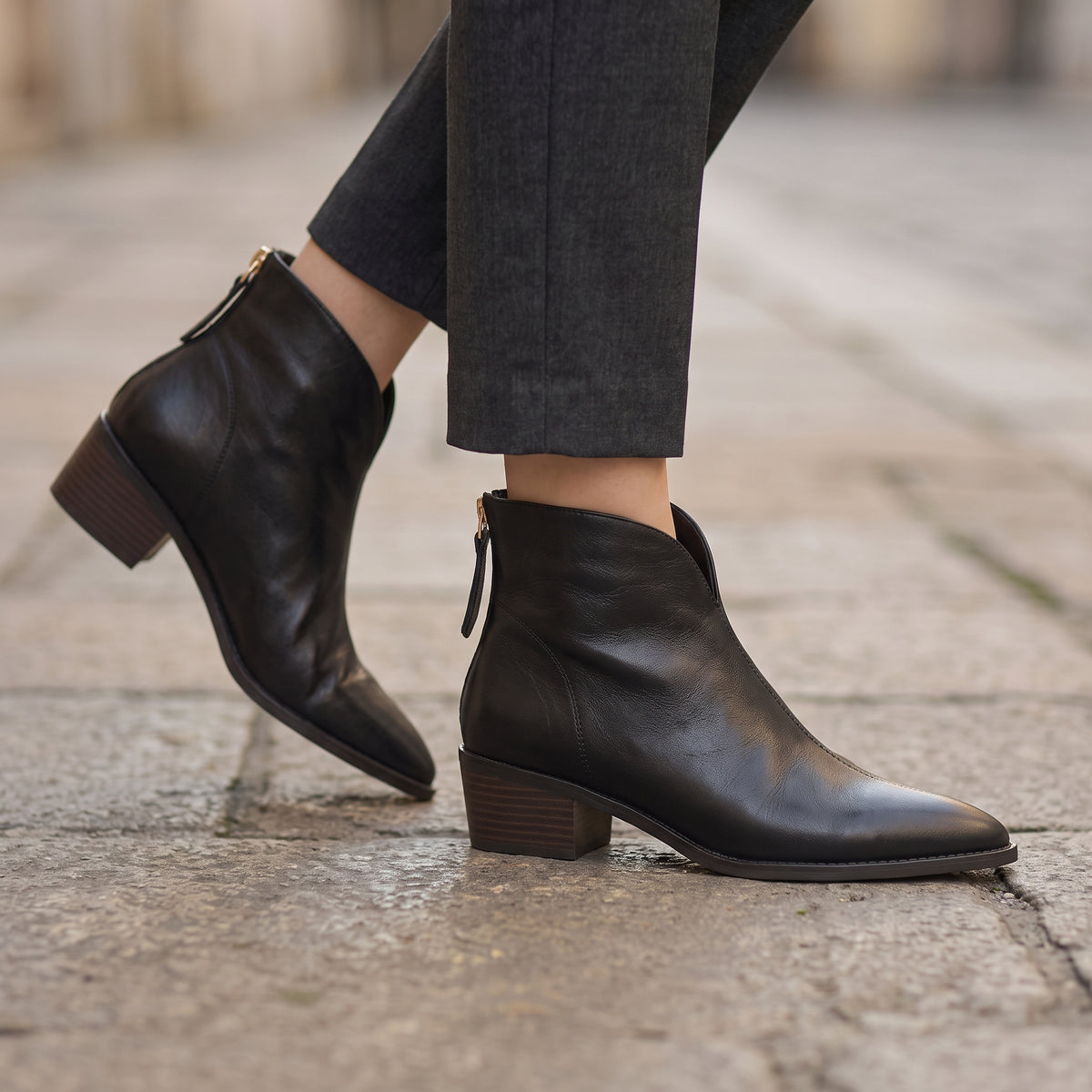 Black leather ankle boots with side zippers and block heels, shown on a model wearing dark gray trousers while walking on a stone pavement.