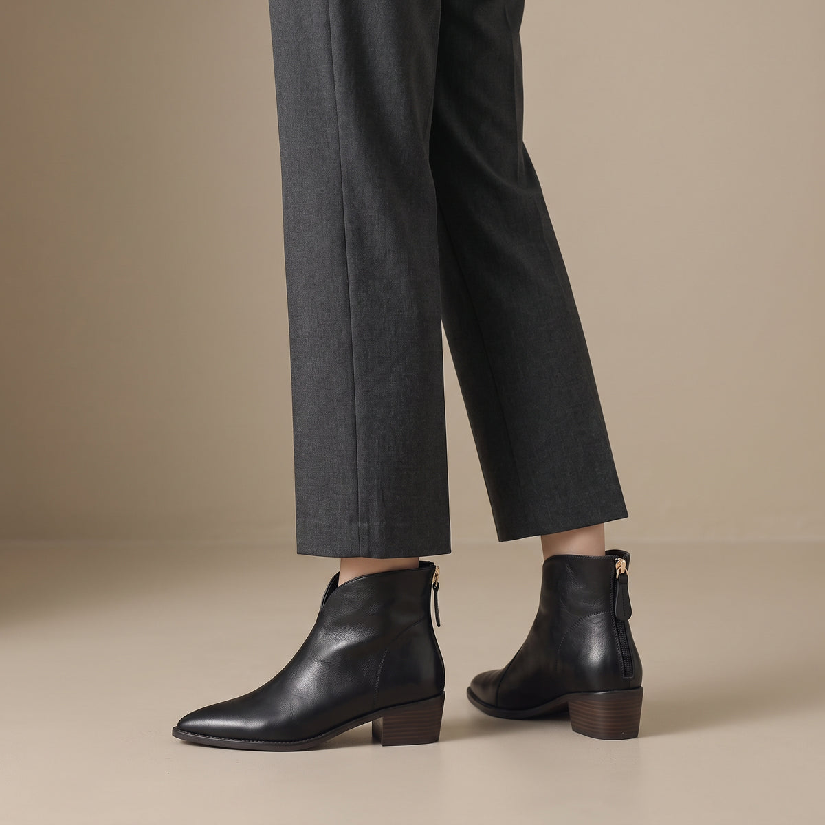Black leather ankle boots with a side zipper and low block heel, shown on a model wearing dark gray trousers against a beige studio background.