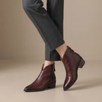 A person wearing dark brown leather ankle boots with pointed toes and low block heels, paired with dark gray trousers, shown against a neutral beige studio background.