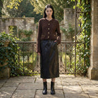 Woman in a brown cardigan and black skirt standing in an outdoor setting with stone walls and iron gates.