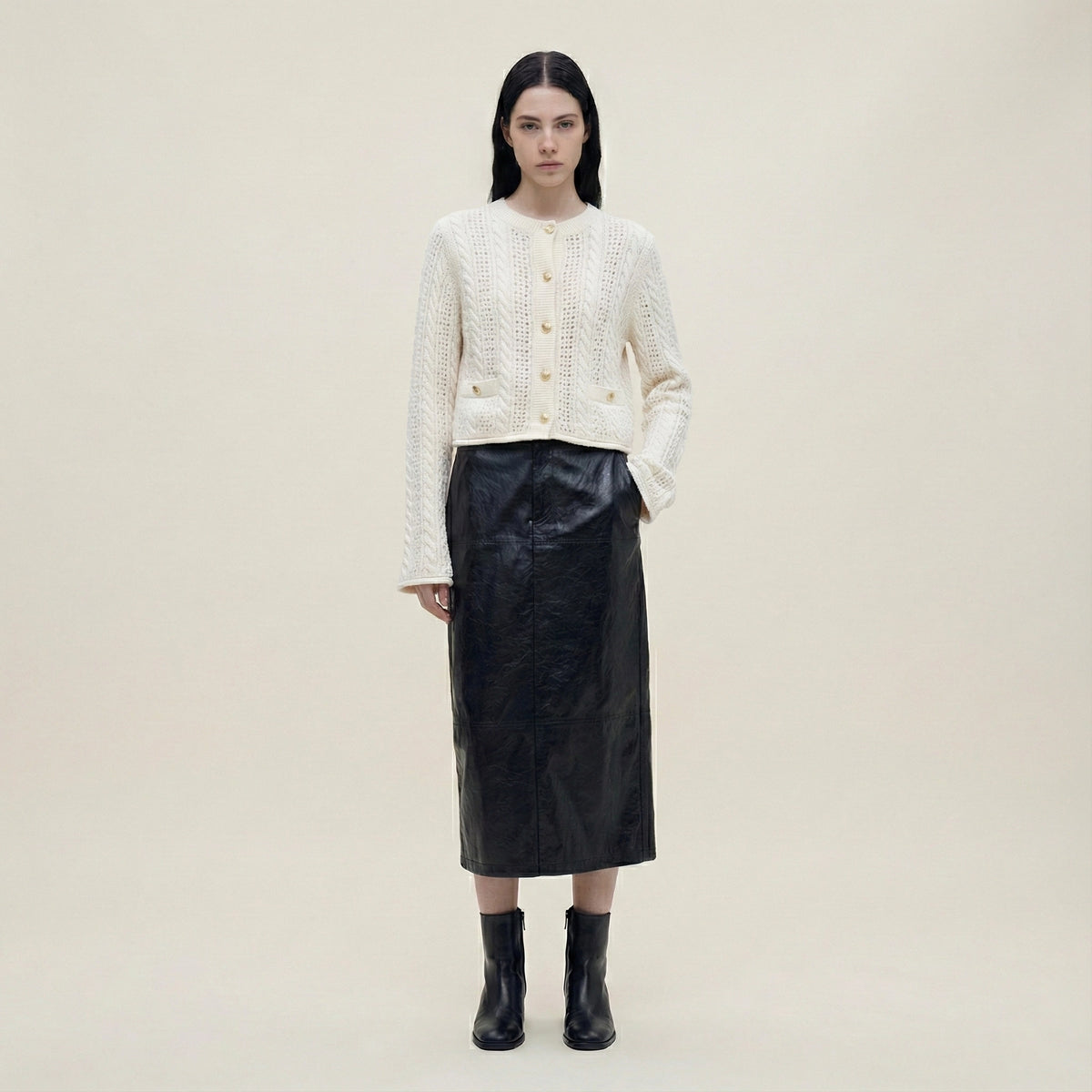 Woman wearing a white textured cardigan, black leather skirt, and black boots on a beige background