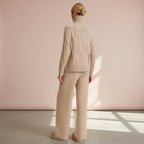 Person wearing a beige cable knit sweater and pants against a pink wall.