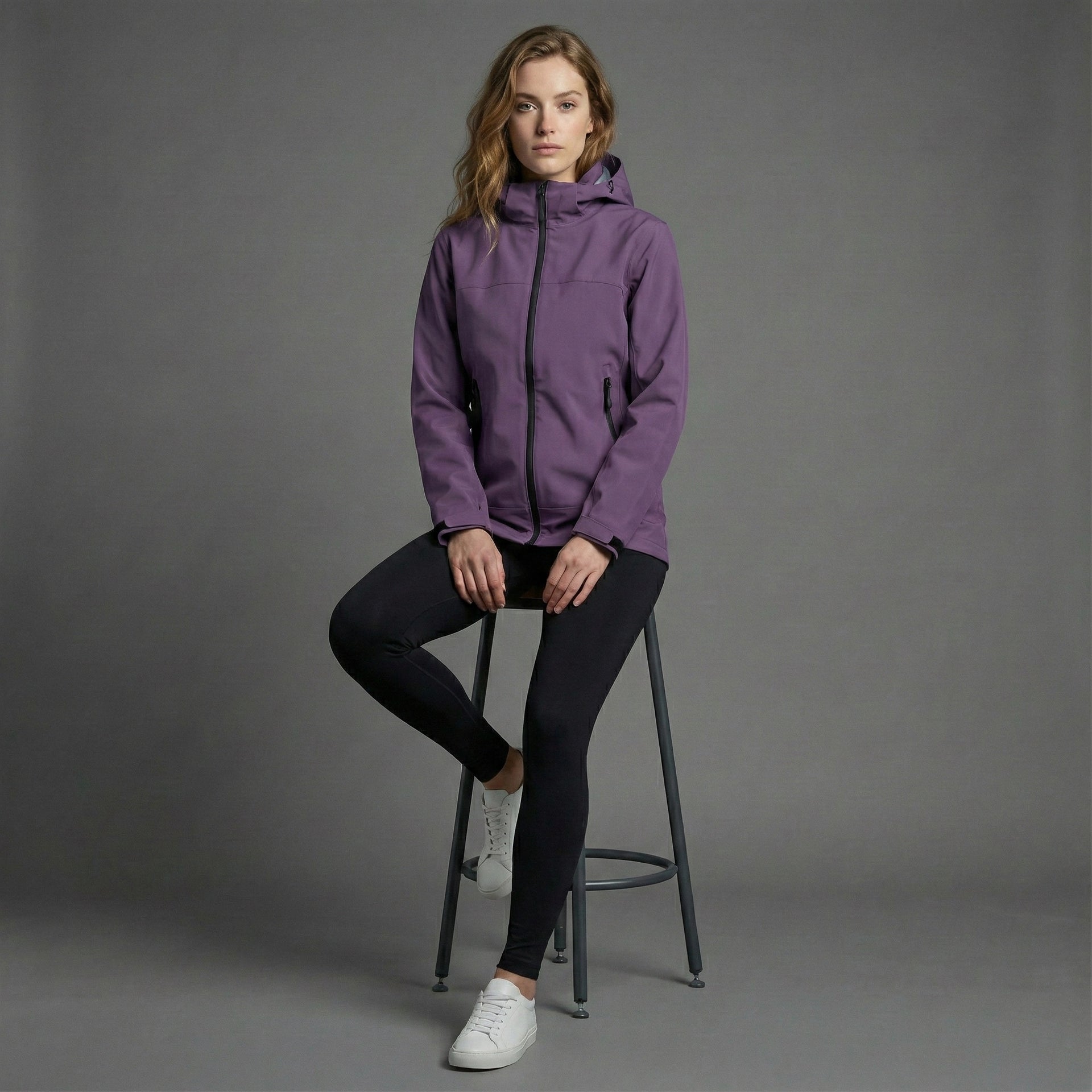 Woman wearing a purple jacket sitting on a stool against a gray background
