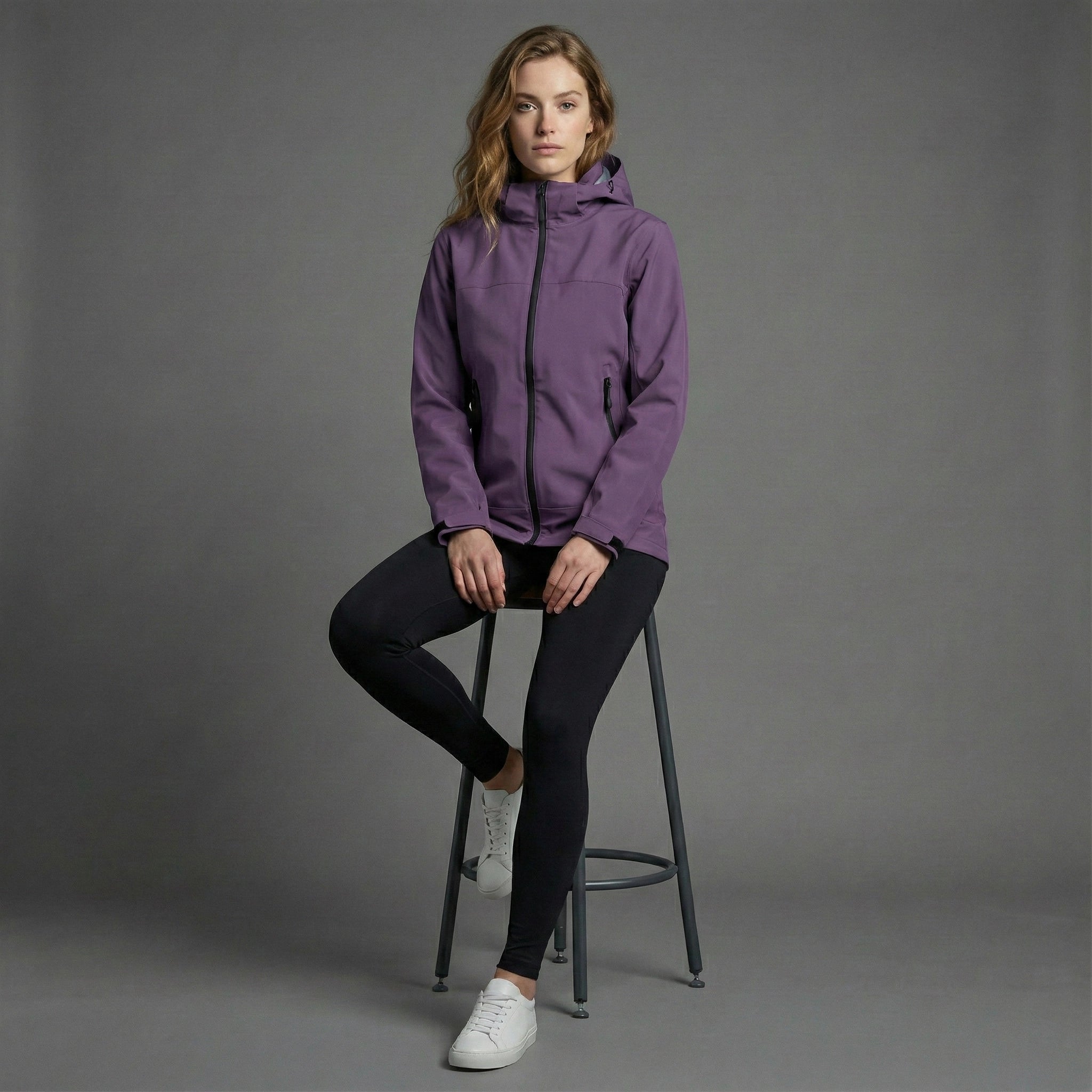 Woman wearing a purple jacket sitting on a stool against a gray background