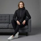 Woman sitting on a gray couch wearing a black jacket and leggings with white sneakers against a gray background