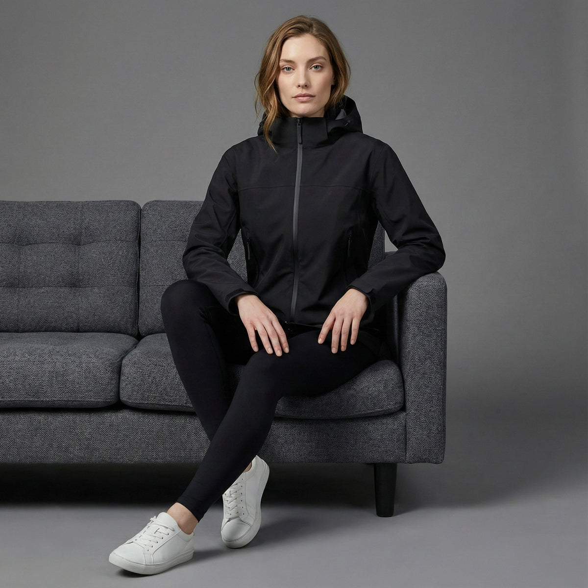 Woman sitting on a gray couch wearing a black jacket and leggings with white sneakers against a gray background