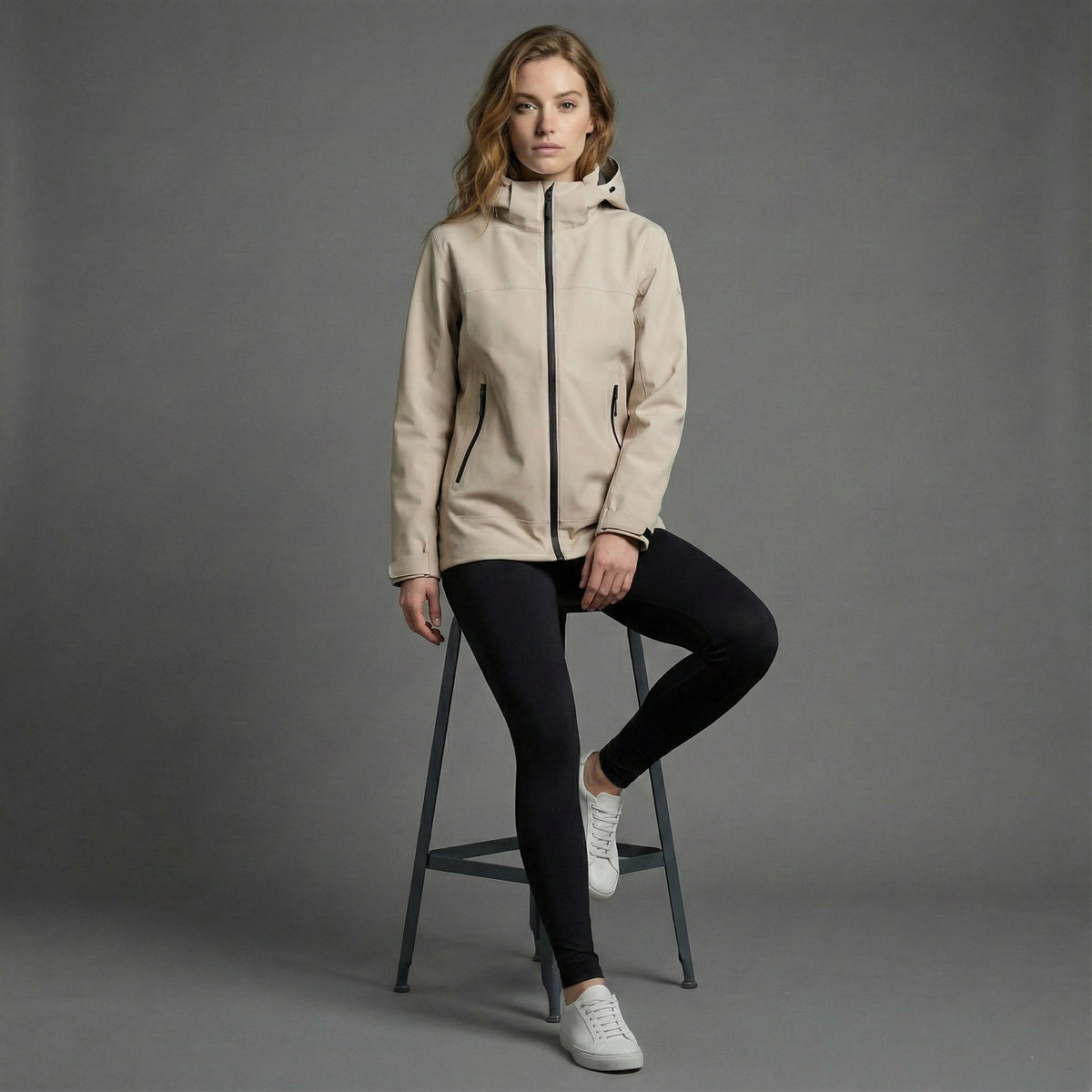 Woman wearing a beige jacket sitting on a stool against a gray background