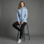Woman wearing a light blue jacket sitting on a stool against a gray background
