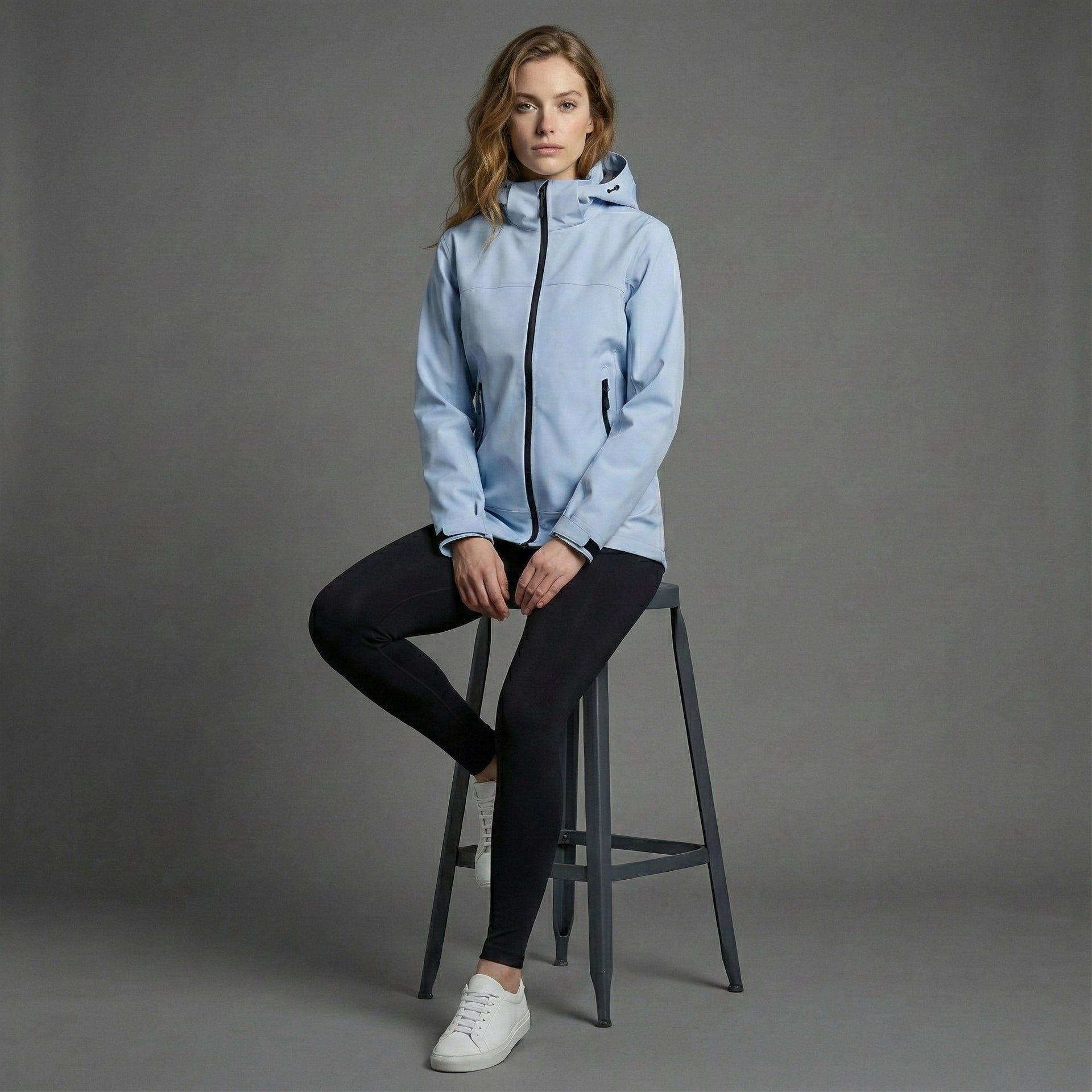 Woman wearing a light blue jacket sitting on a stool against a gray background