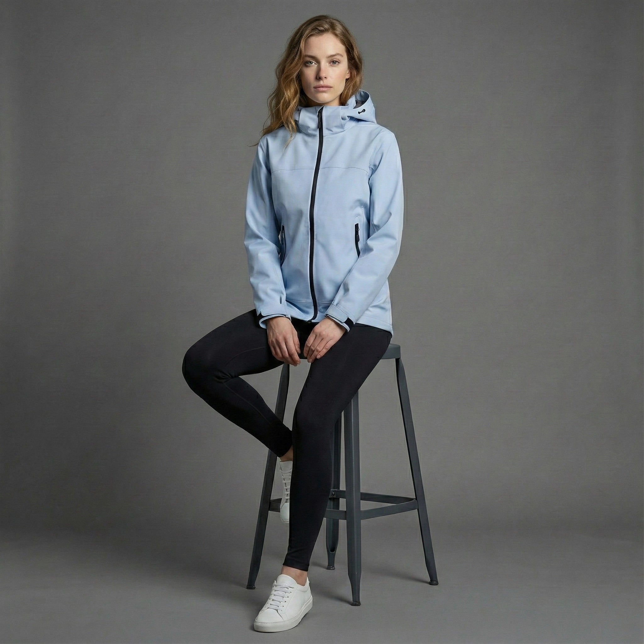 Woman wearing a light blue jacket sitting on a stool against a gray background