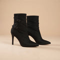 A pair of black suede ankle boots with pointed toes, slim high stiletto heels, and decorative wraparound straps with silver-tone buckles, set against a solid beige background.
