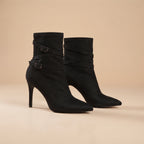 A pair of black suede ankle boots with pointed toes, slim high stiletto heels, and decorative wraparound straps with silver-tone buckles, set against a solid beige background.