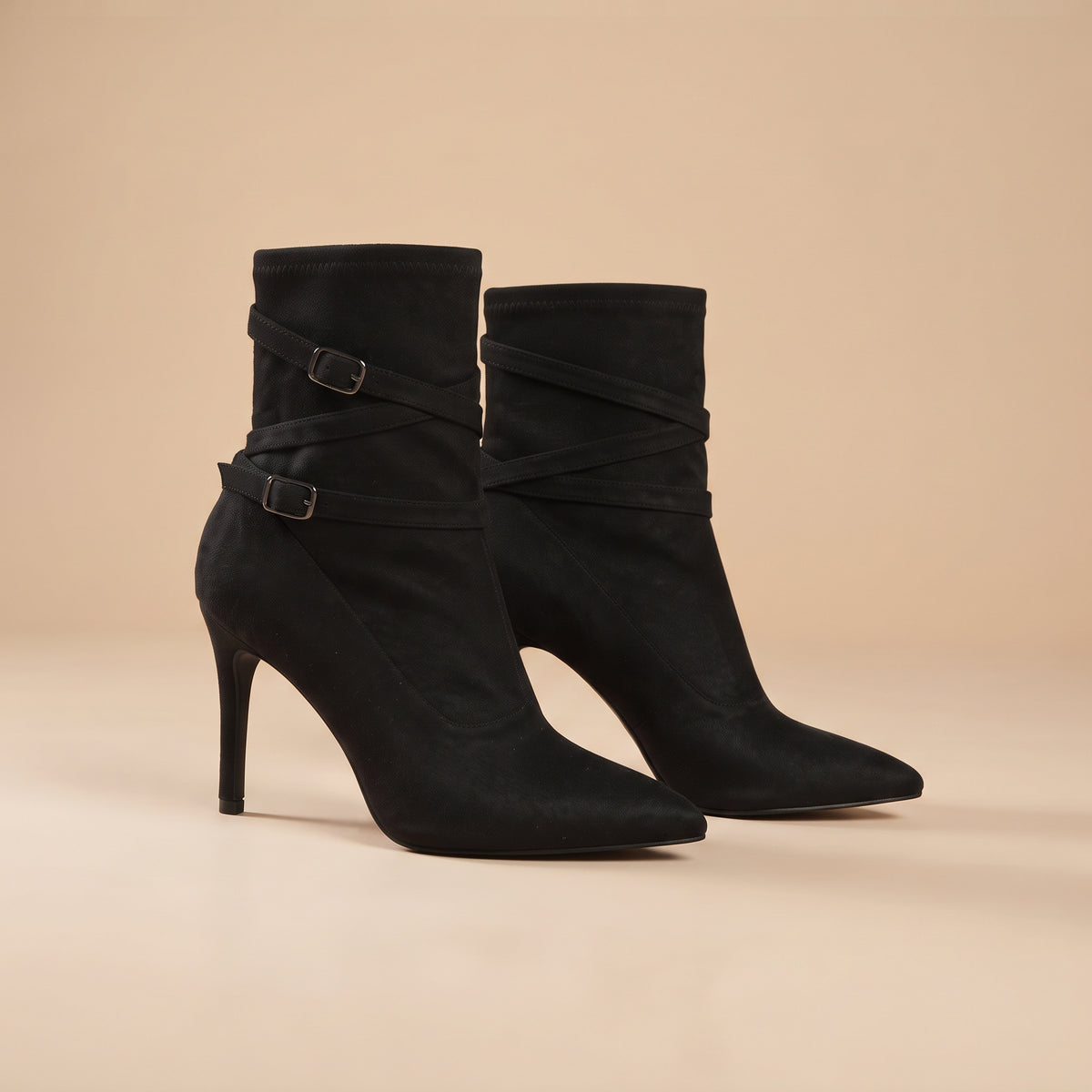 A pair of black suede ankle boots with pointed toes, slim high stiletto heels, and decorative wraparound straps with silver-tone buckles, set against a solid beige background.