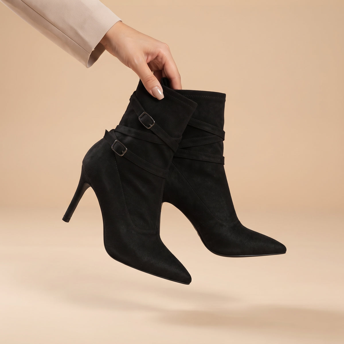 Black ankle boots with pointed toes, high stiletto heels, and double buckle straps, held by a hand wearing a beige sleeve against a warm beige studio background.