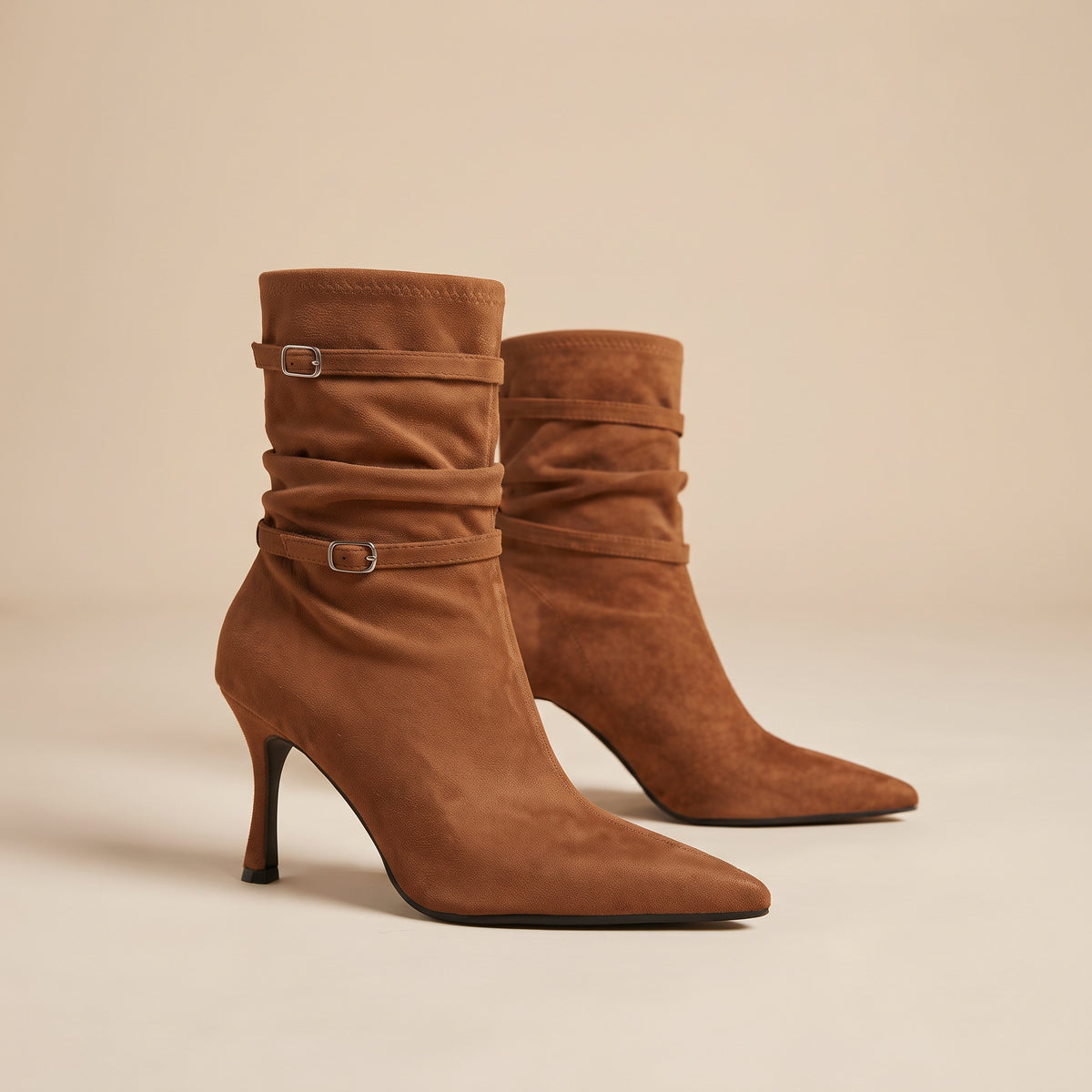 A pair of brown suede ankle boots with pointed toes, slim high heels, and decorative buckle straps around the shaft, photographed on a clean beige studio background.