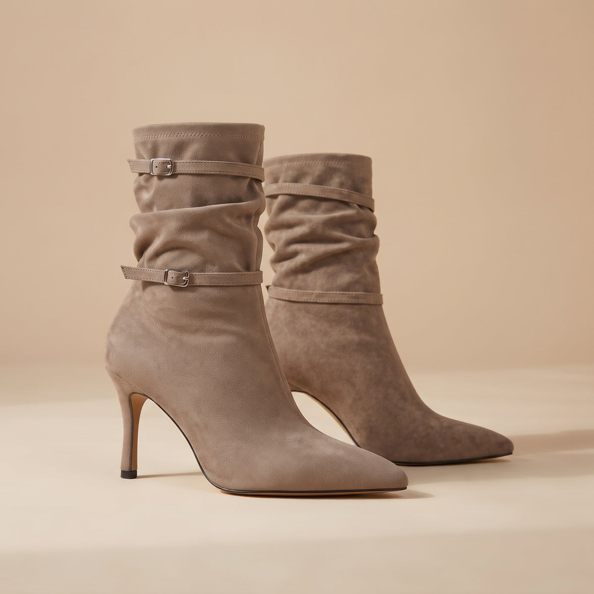 A pair of taupe suede ankle boots with pointed toes, slim high stiletto heels, and decorative buckle straps around the shaft, set against a warm beige background.