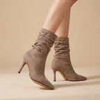 A pair of taupe-brown suede slouchy ankle boots with kitten heels and buckle straps, shown on a model's legs against a warm beige studio background.