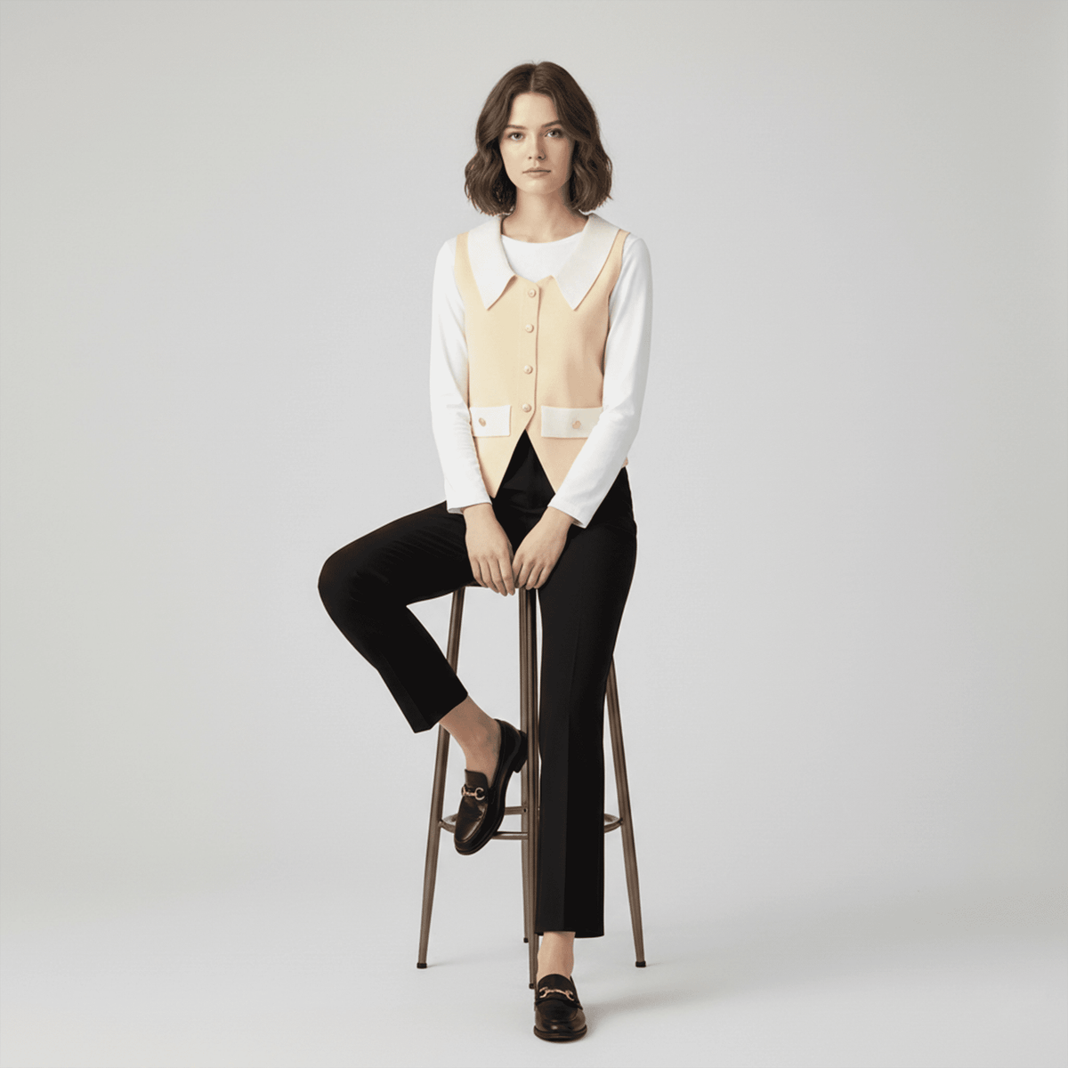 Woman sitting on a stool wearing a white blouse, beige vest, black pants, and black shoes against a plain background.
