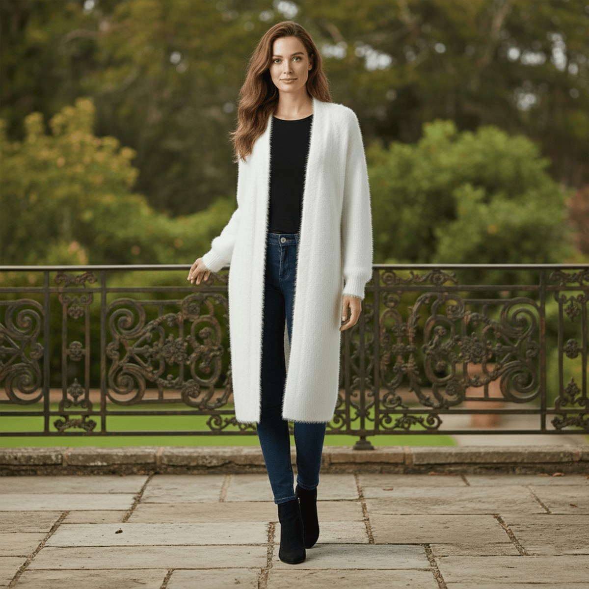 Woman wearing a long white coat standing in a park-like setting with greenery and a decorative metal fence.