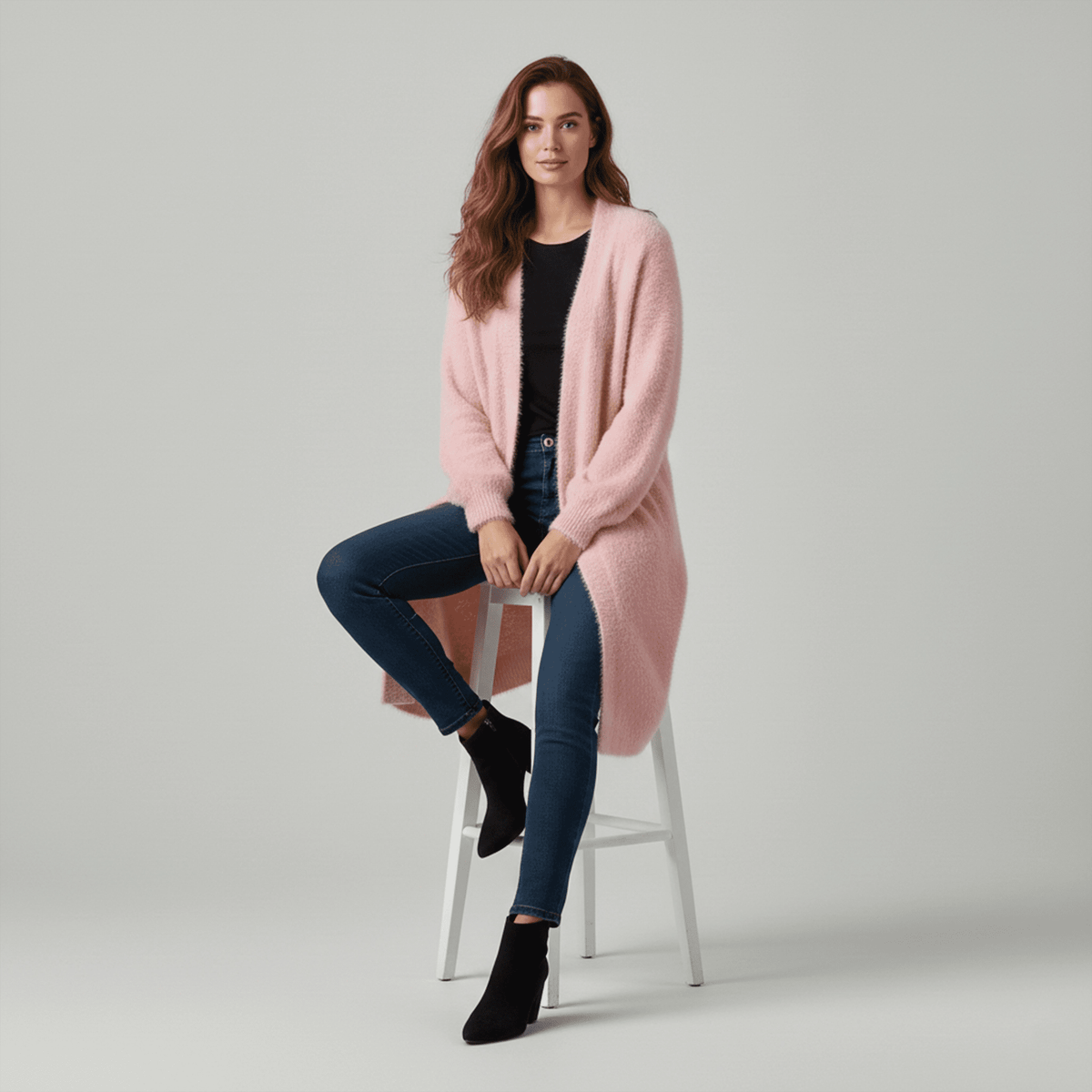 Woman wearing a pink cardigan, black top, and jeans sitting on a stool against a plain background