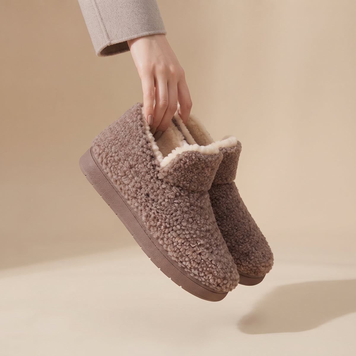 A pair of cozy ankle-height slipper boots in a textured taupe-brown sherpa fabric with a cream lining, held by a hand wearing a light gray sleeve against a warm beige background.