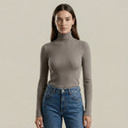 Woman wearing a gray turtleneck sweater and blue jeans against a beige background