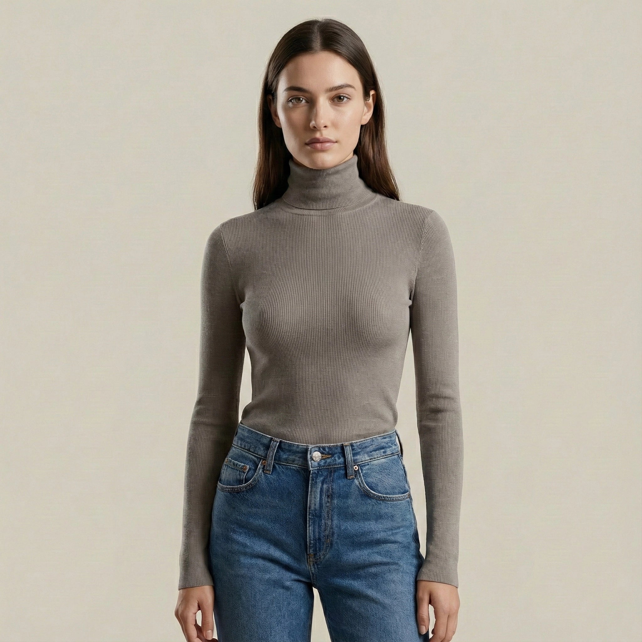 Woman wearing a gray turtleneck sweater and blue jeans against a beige background