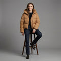 Person wearing a brown puffer jacket sitting on a wooden stool against a gray background