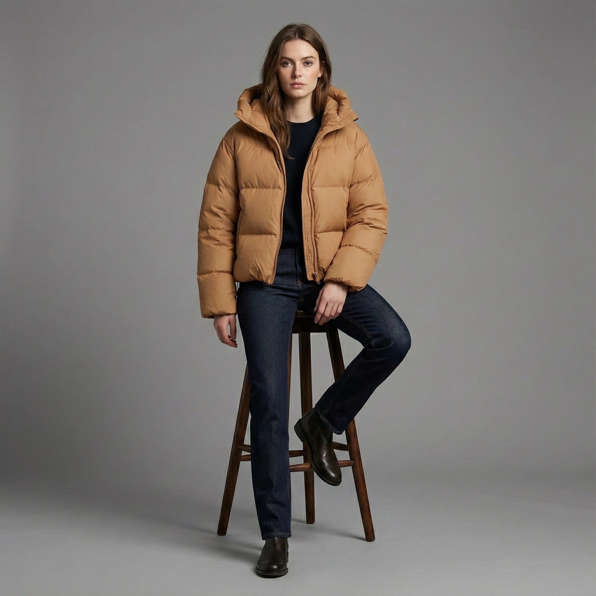 Person wearing a brown puffer jacket sitting on a wooden stool against a gray background