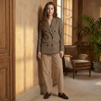 Woman in a checkered blazer and beige pants standing in a sunlit room with wooden furniture and a plant.