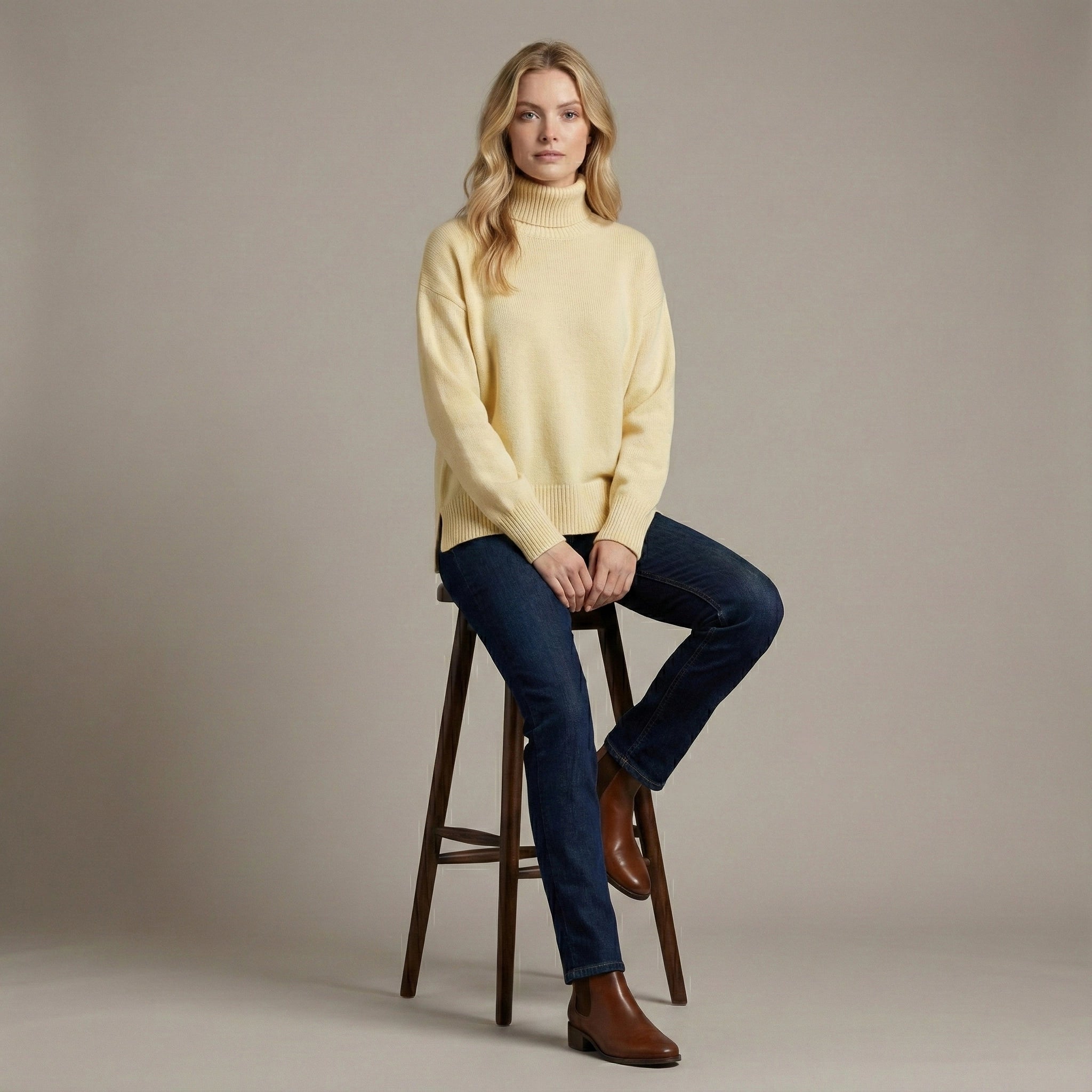 Woman wearing a yellow sweater and blue jeans sitting on a wooden stool against a plain background