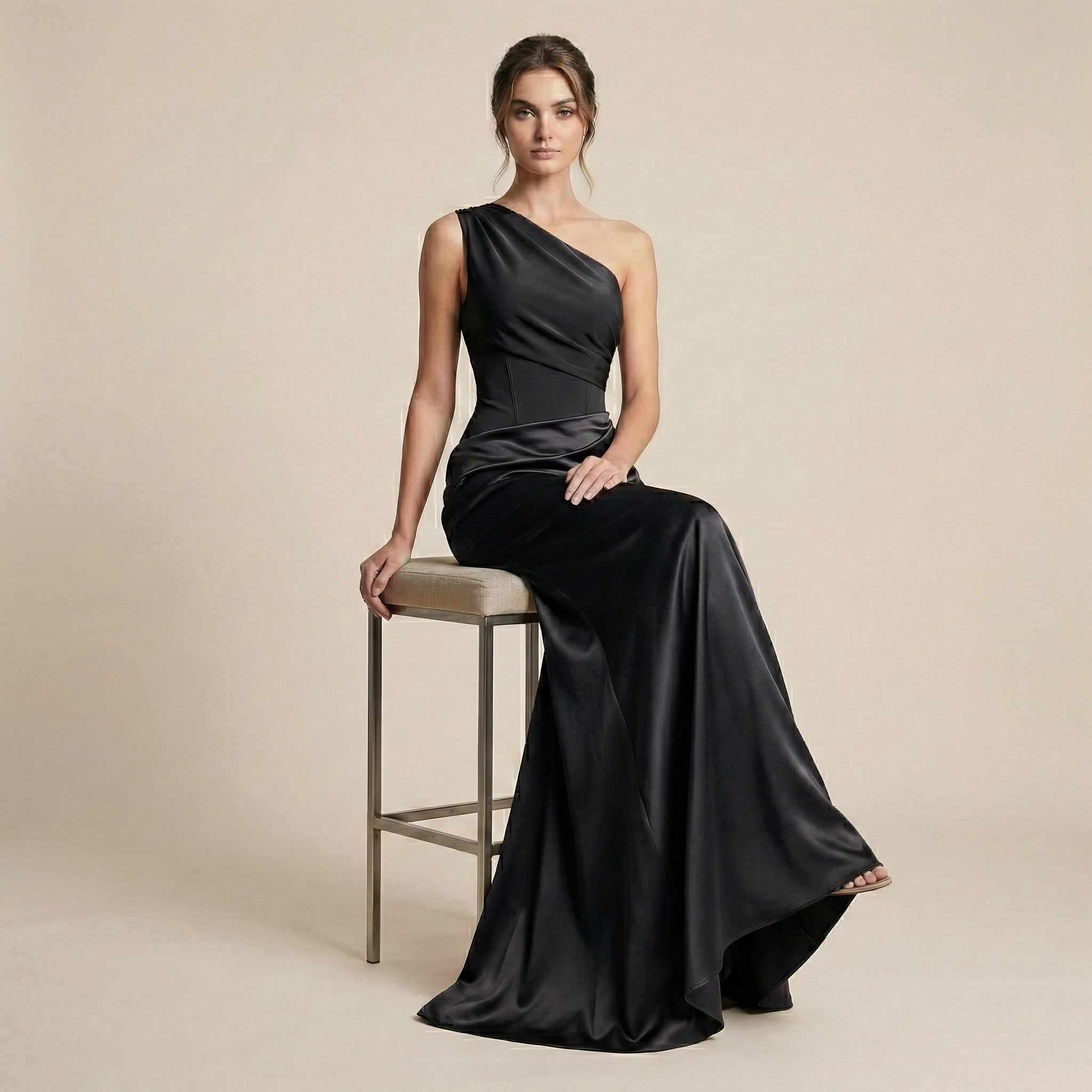 Woman in a black one-shoulder gown sitting on a stool against a beige background