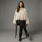 Woman sitting on a chair wearing a beige shirt and black pants against a gray background