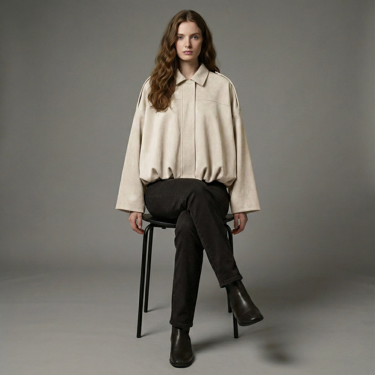 Woman sitting on a chair wearing a beige shirt and black pants against a gray background