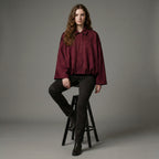 Woman wearing a burgundy shirt and black pants sitting on a stool against a gray background