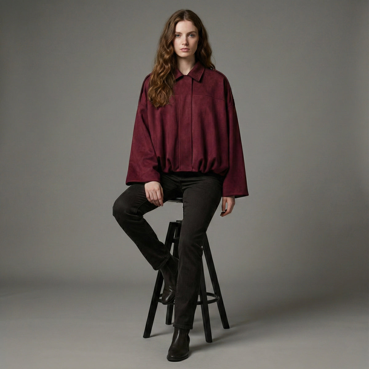 Woman wearing a burgundy shirt and black pants sitting on a stool against a gray background