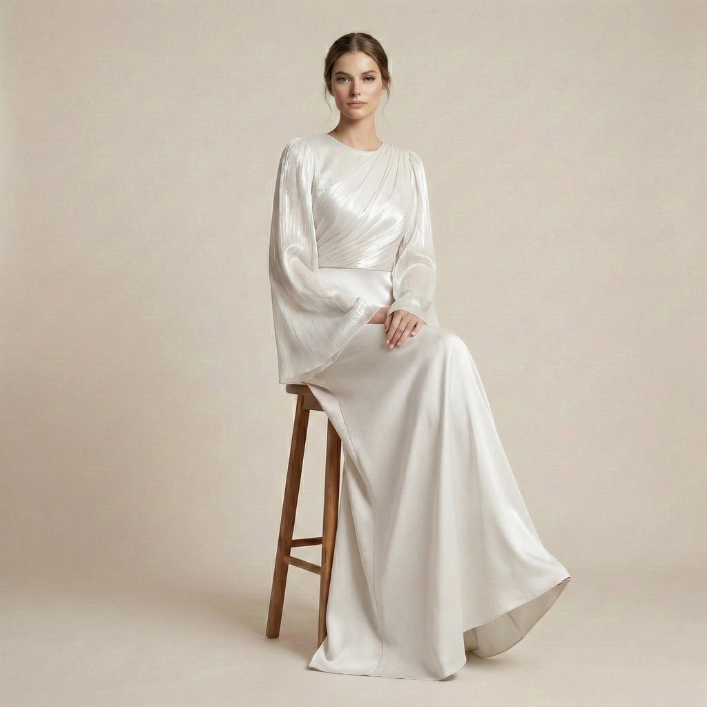 Woman in a long, white dress sitting on a wooden stool against a beige background
