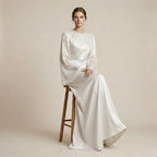 Woman in a long, white dress sitting on a wooden stool against a beige background