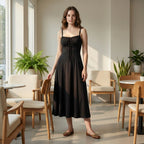 Woman in a black dress standing in a modern dining area with wooden chairs and plants.
