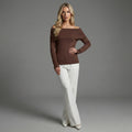 Woman wearing a brown off-shoulder top and white pants against a gray background