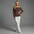 Woman wearing a brown off-shoulder top and white pants against a gray background