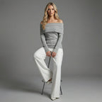 Woman wearing a gray off-shoulder top and white pants sitting on a stool against a gray background