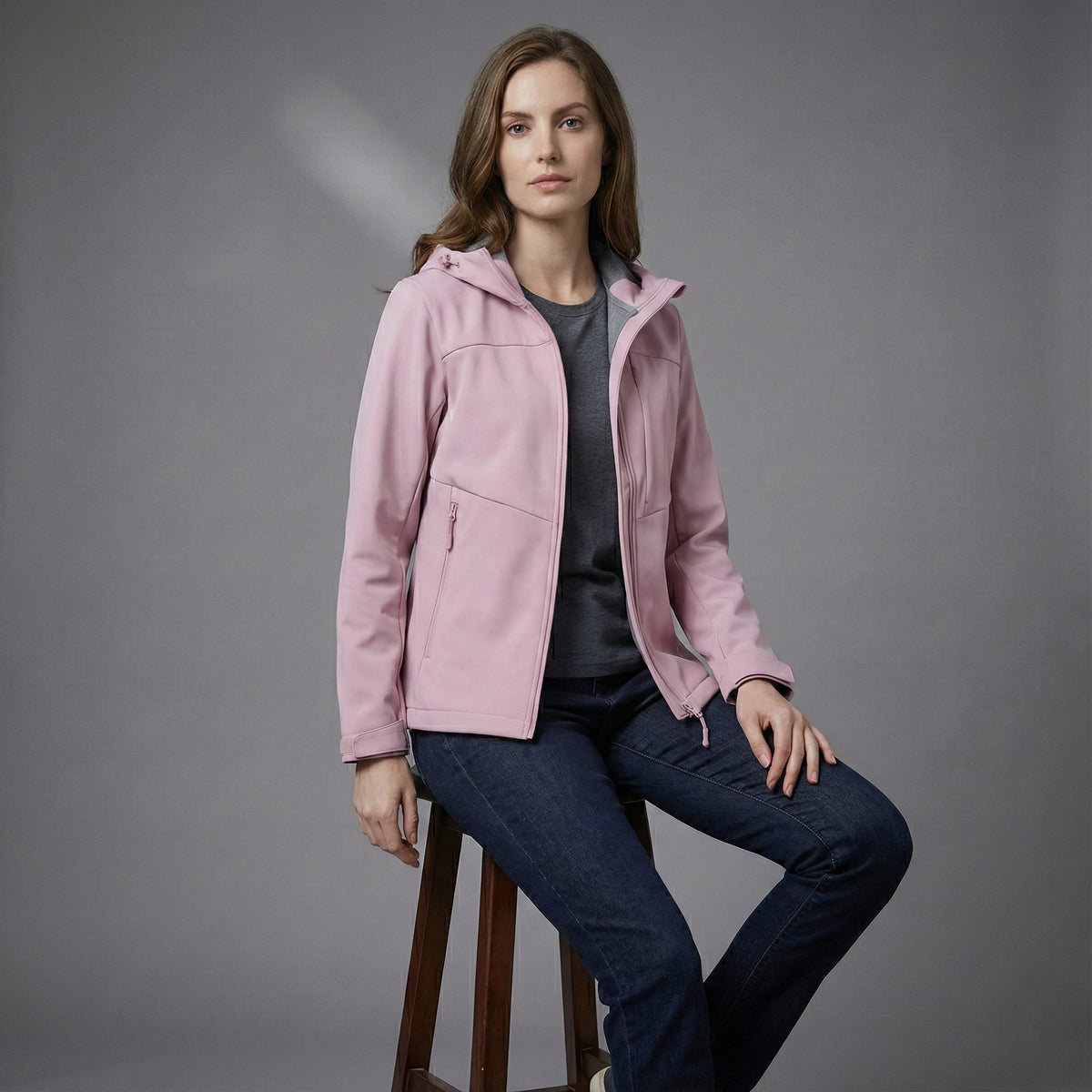 Woman wearing a pink jacket sitting on a stool against a gray background