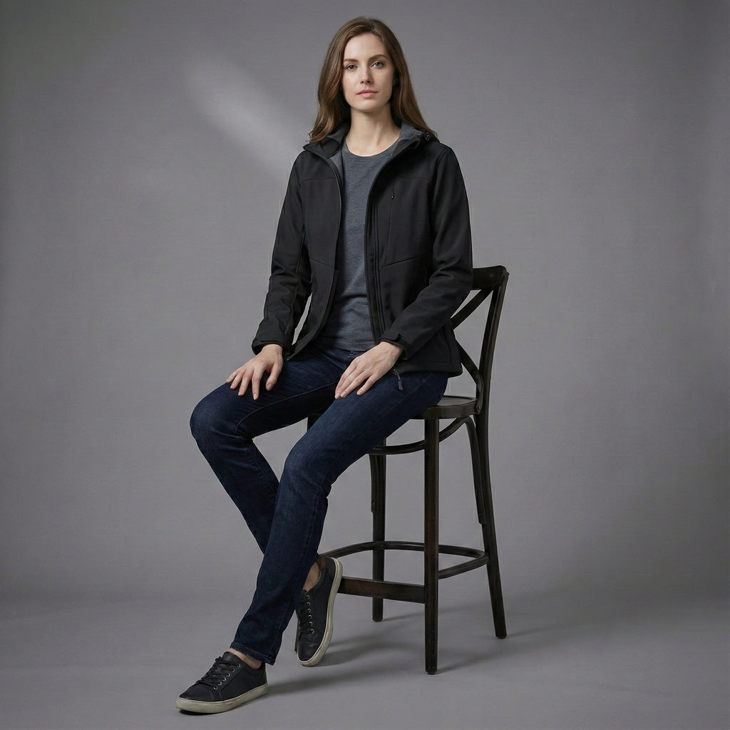 Woman sitting on a chair wearing a black jacket and blue jeans against a gray background