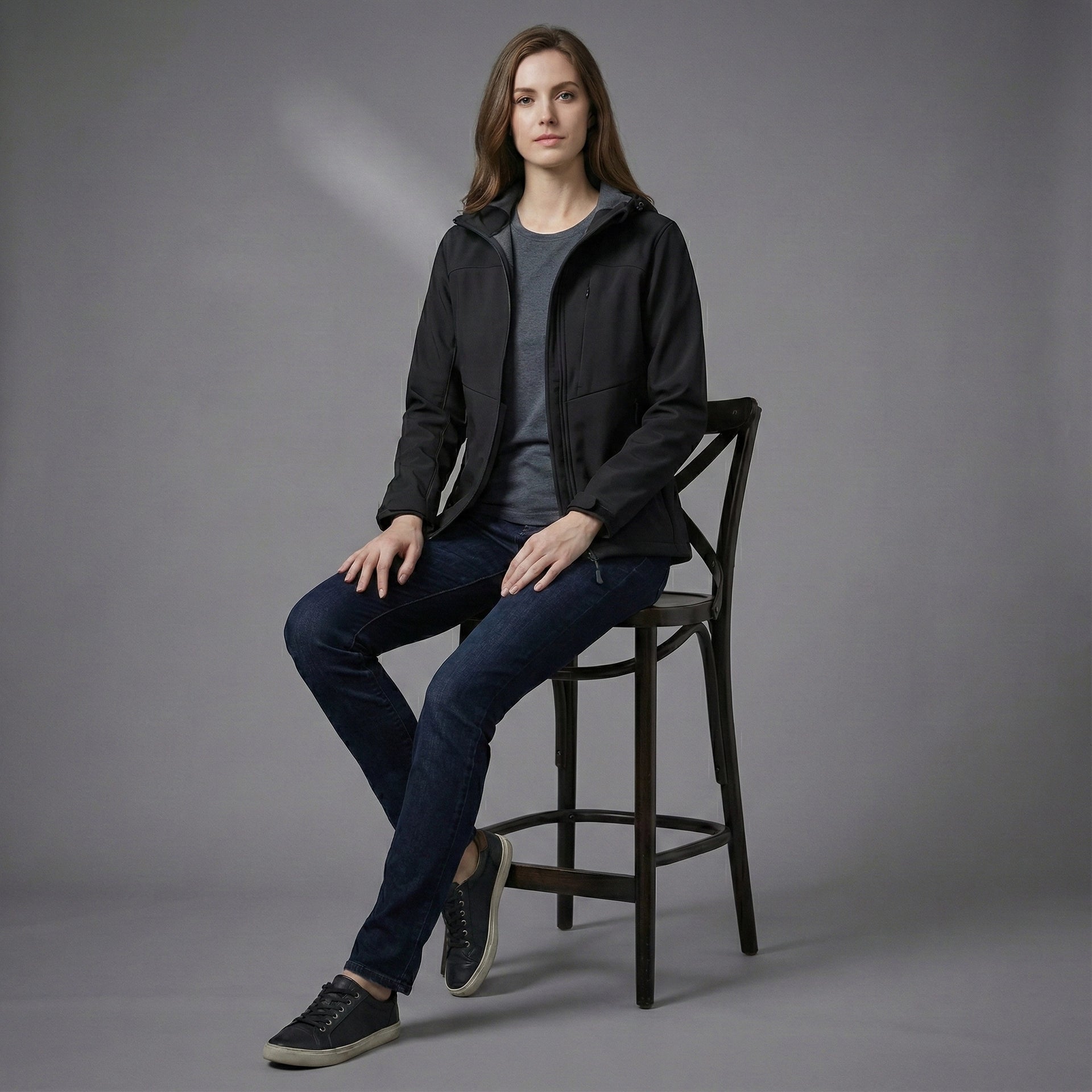 Woman sitting on a chair wearing a black jacket and blue jeans against a gray background