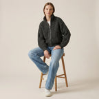 Person wearing a dark gray zip-up sweater and light blue jeans sitting on a wooden stool against a beige background.