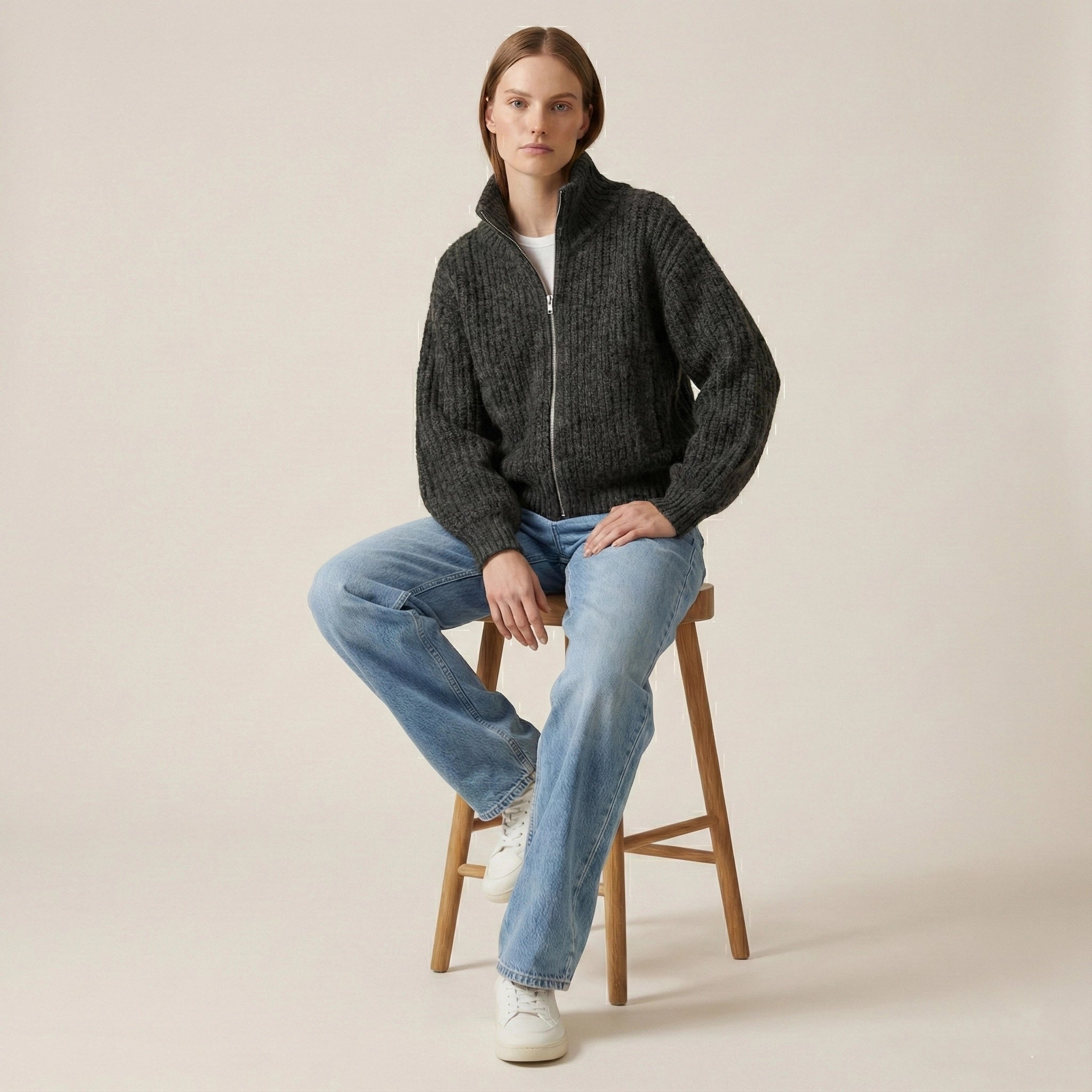 Person wearing a dark gray zip-up sweater and light blue jeans sitting on a wooden stool against a beige background.